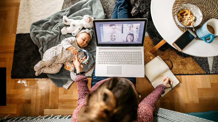 Mom on video call with her team, taking notes and engaging with her baby.